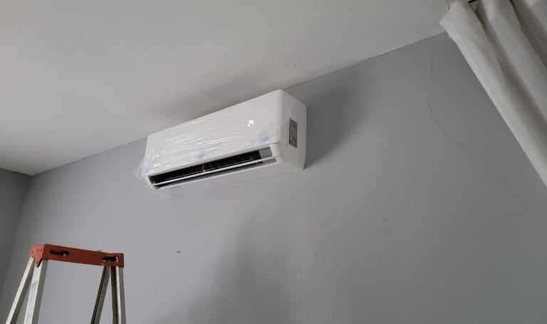 A wall-mounted ductless mini split air conditioner unit being installed on a grey wall with a stepladder nearby.