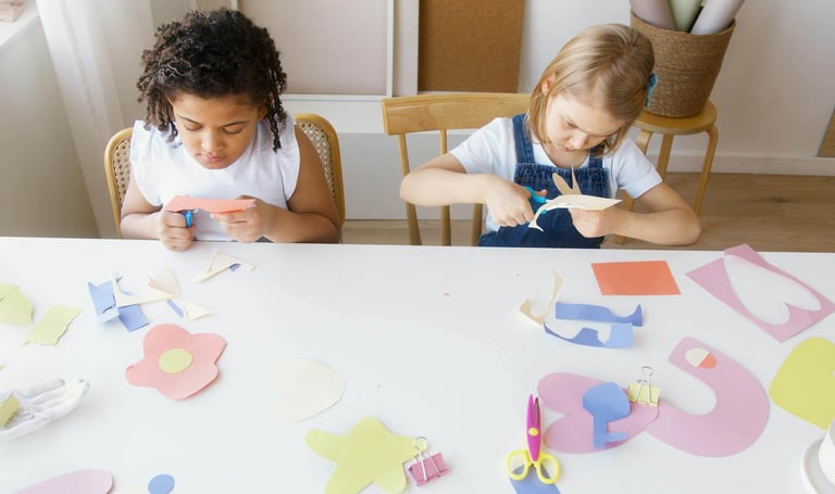 Two young girls cutting paper with scissors whilst making crafts at a table.