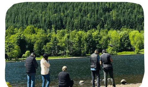 a group of people standing beside a loch with trees across the water