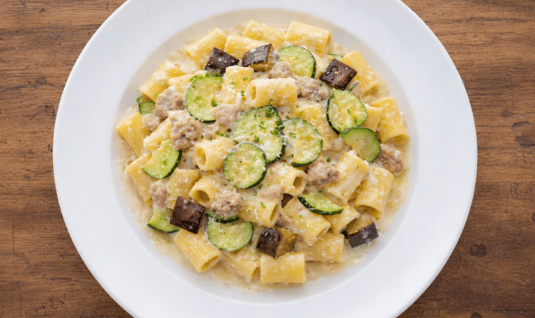 Rigatoni pasta with zucchini, eggplant, and sausage in a creamy sauce on a white plate.