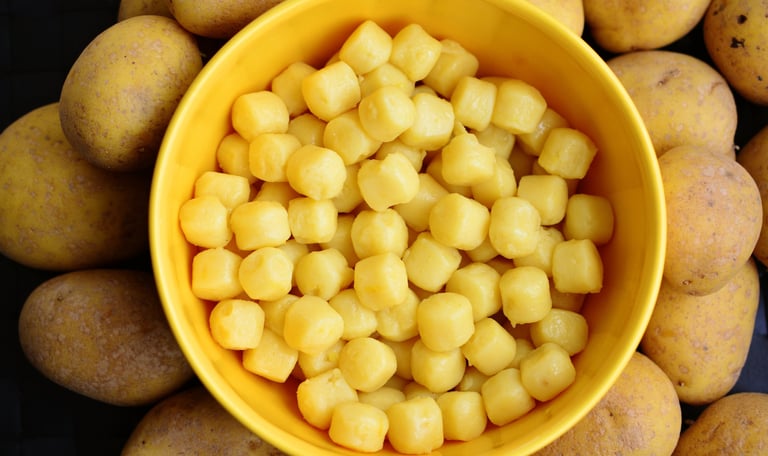 Fresh homemade potato gnocchi served in a bright yellow bowl surrounded by raw whole potatoes.
