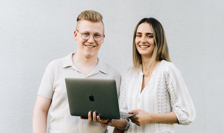 Raphael und Vanessa Bogner stehen vor einer weißen Wand mit einem Macbook in der Hand