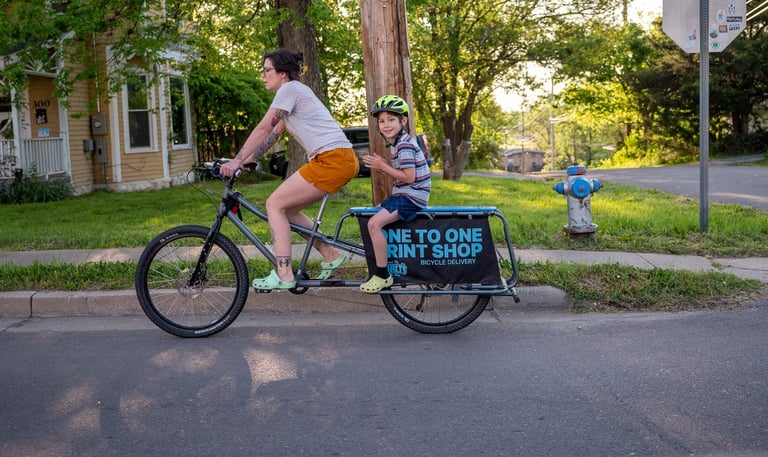 A women riding a bike with a young boy sitting on the back rack with a sign that reads "One to One"