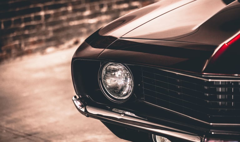 Close-up of a classic black muscle car headlight and chrome bumper with a vintage brick background.