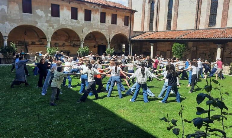 Persone in cerchio su prato, si tengono per mano e danzano in Museo