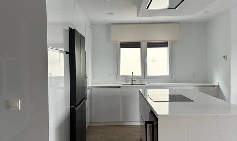 Cocina blanca, isla, electrodomésticos negros, suelo madera.