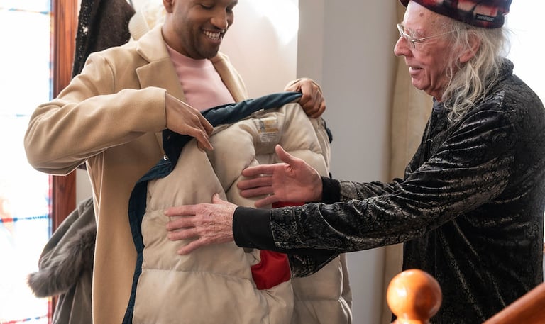 An older man helps a younger man try on a tan winter coat inside a brightly lit home foyer.
