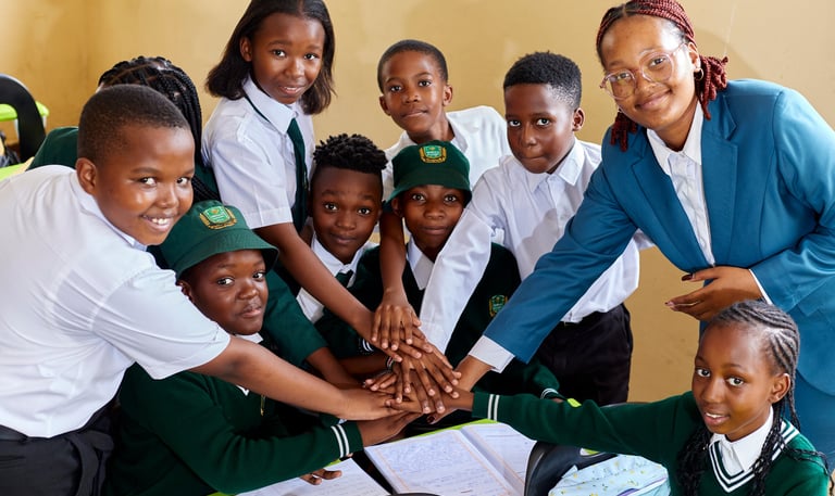 Diverse African students in school uniforms and their teacher stack hands in a classroom team huddle.