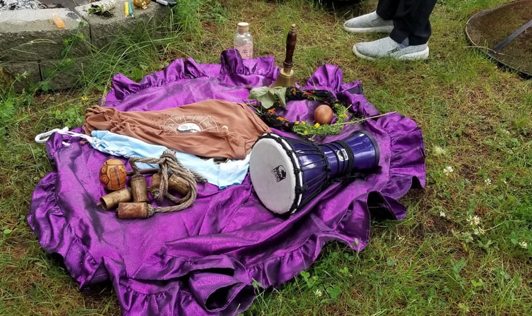 Outdoor ceremonial setup on a purple cloth with a drum, bells, moon water, zodiac cloth, and ritual