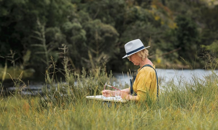 A woman artist wearing a hat paints on a canvas while sitting in a grassy field by a river.