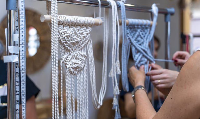 Mujeres anudando nudos en tejido de macramé con hilo color azul claro que cuelga de un palo de madera
