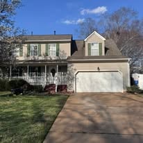 Two-story tan suburban house with a white garage door, front porch, and green shutters.