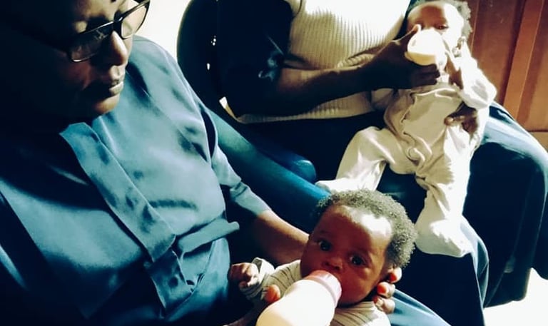 African Catholic nuns bottle-feeding two young infants at a childcare mission or orphanage center.