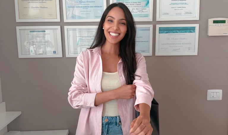 A smiling female receptionist in a pink shirt standing at a dental clinic desk with medical diplomas on the wall.