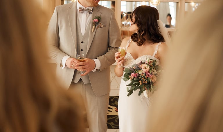 A bride and groom in a light grey suit holding drinks and flowers at their indoor wedding reception.