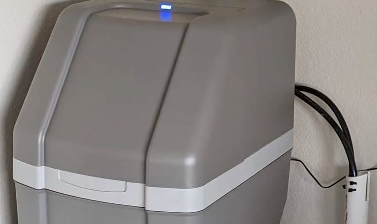 A gray Rheem water softener unit installed against a wall with stainless steel flexible pipes.