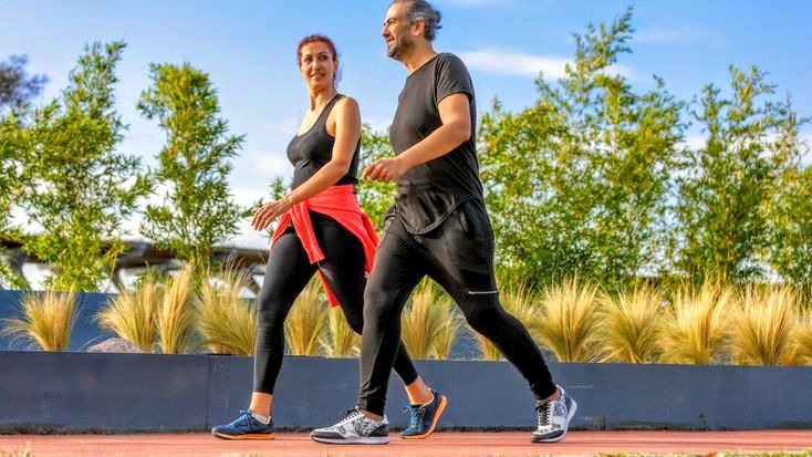 Casal caminhando ao ar livre em pista cercada por plantas, em dia ensolarado.