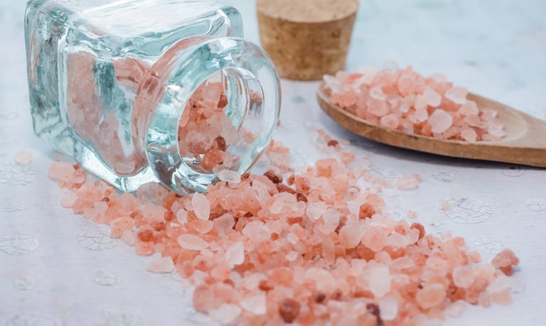 Pink Himalayan salt spilled from a glass jar onto a surface with a wooden spoon nearby.