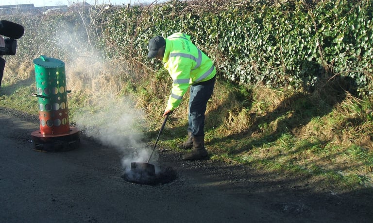 A worker in high-visibility gear repairs a pothole on a rural road using steaming asphalt.