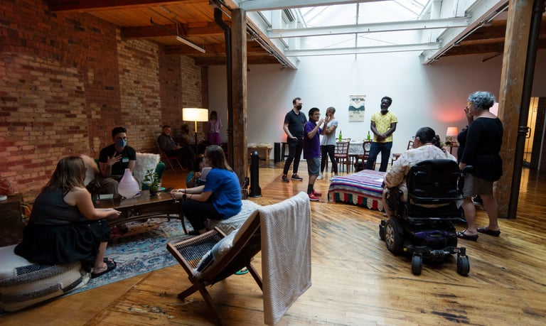 Gallery view of a living room with a bed, poufs, carpet, and about 20 people in the space.