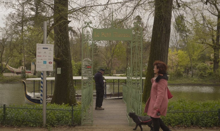 A woman walks her dog past the Le Petit Mousse gate at a scenic park with a canal and boat.