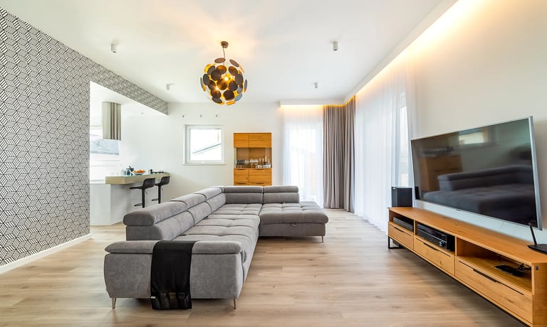 Nowoczesny salon z szarą narożną sofą, telewizorem na drewnianej szafce i aneksem kuchennym.