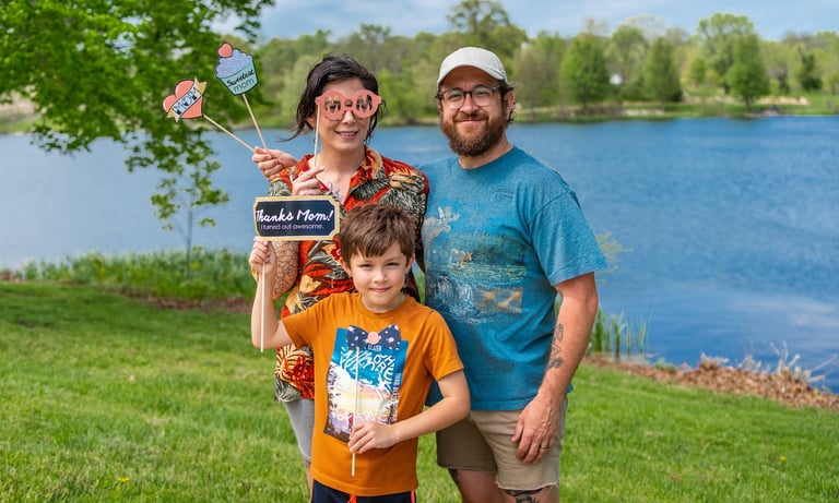 Staged photo of a couple with a young child in front of a lake, holding up paper props