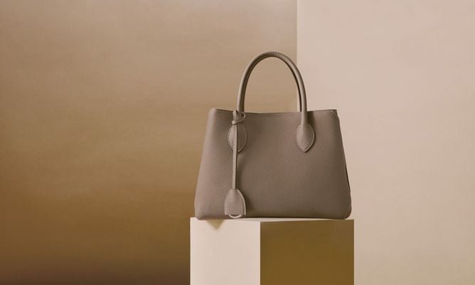 Elegante bolso de mano de piel color topo exhibido sobre un pedestal minimalista.