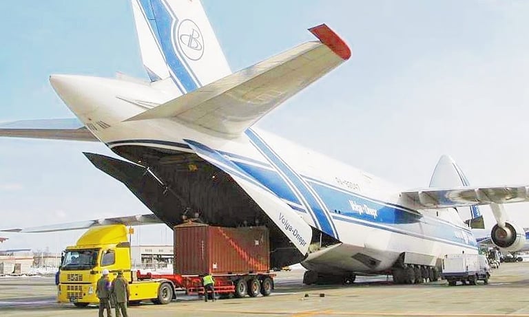 A Volga-Dnepr Antonov An-124 cargo plane loading a shipping container from a yellow truck at an airport.