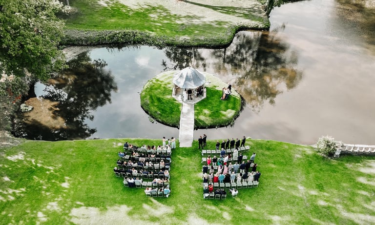 Drone photo of an outdoor wedding ceremony by a lakeside gazebo surrounded by guests