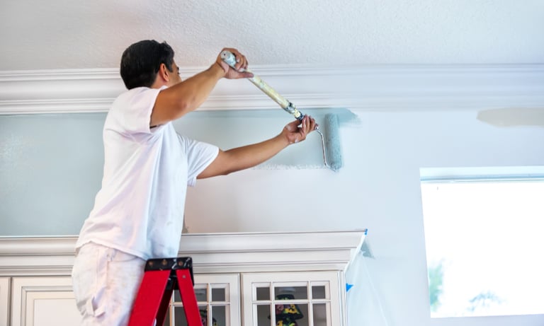 Professional house painter on a ladder using a roller to apply light blue paint to an interior wall.