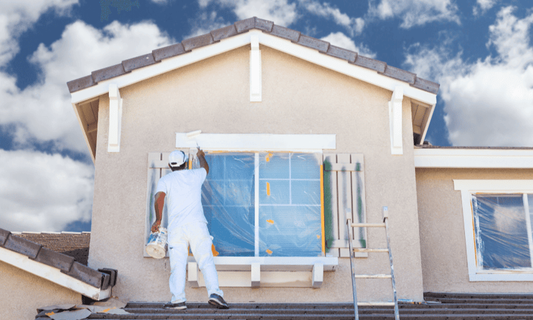 Professional house painter applying white paint to a home exterior window trim under a blue sky.