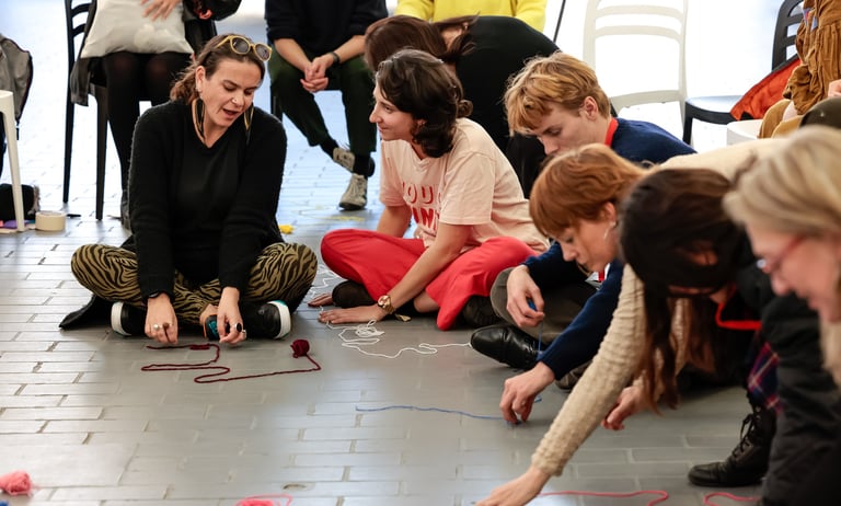 Eight adults sitting on the ground making patterns with colourful yarn.