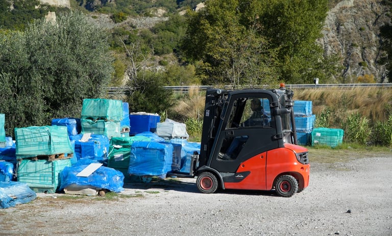 Red and black forklift moving blue plastic-wrapped pallets at an outdoor industrial site.