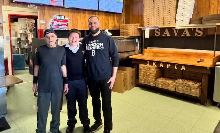 Three men smiling inside Sava's Pizza shop with stacked pizza boxes and menu boards.