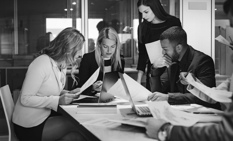 a group of people sitting around a table looking intently at paperwork and screens
