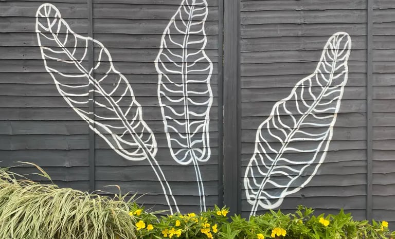 Three large banana leaves painted in a modern graphic style on a grey yard fence and flowers.