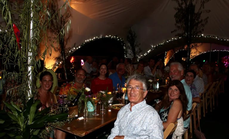 Guests enjoying an outdoor dinner party under a decorated tent with string lights and greenery.