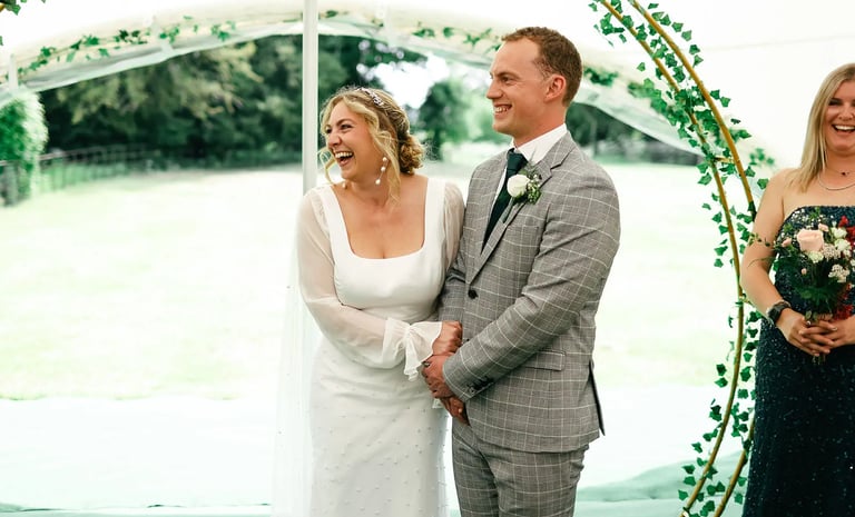 A laughing bride and groom stand together during an outdoor garden wedding ceremony.