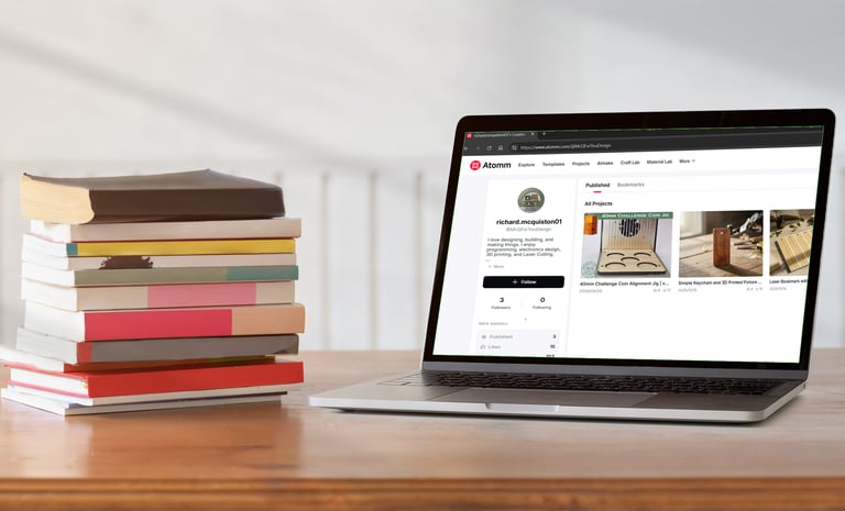 A stack of hardcover books sits next to an open laptop on a wooden desk showing a design website.