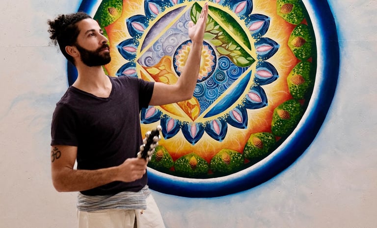 Bearded man in a yoga studio holding bells in front of a colorful sacred geometry mandala wall mural.