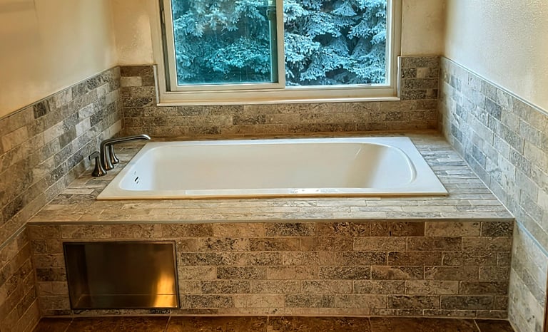 A modern white soaking tub surrounded by grey stone tile and a window overlooking snow-covered pine trees.