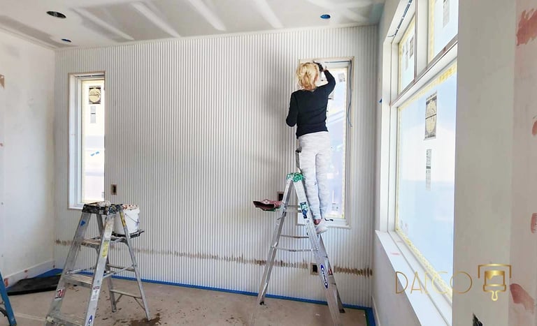 A woman on a ladder paints white textured wall panels during a modern home interior renovation project.