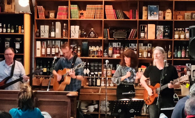 A live band performs folk music in front of a rustic bar shelf filled with vintage books and liquor bottles.