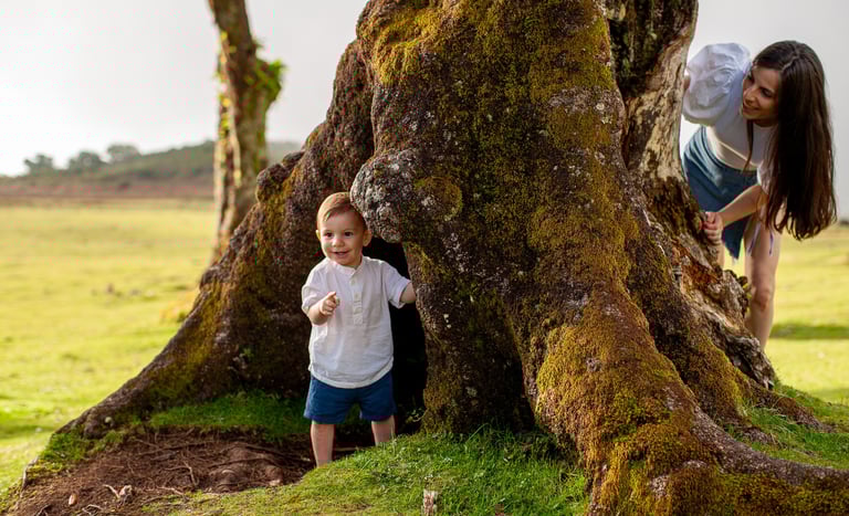 Mother watching toddler son explore around ancient tree trunk during natural family PHOTOSHOOT