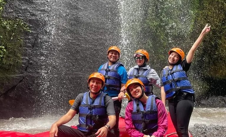 A group of friends in helmets and life jackets posing in a red white-water raft near a waterfall.