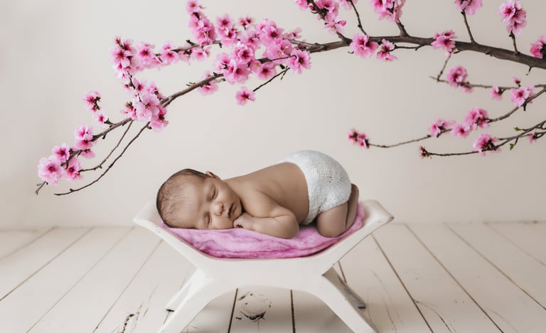 A newborn baby sleeping on a white stool under pink cherry blossom branches for a professional studio photoshoot.