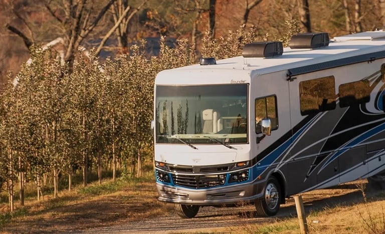 A white Fleetwood Bounder Class A motorhome driving through a scenic rural apple orchard at sunset.