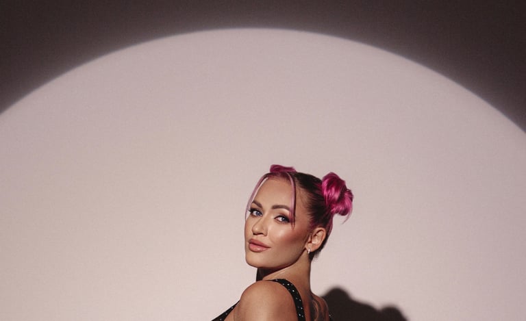 A stylish woman with pink space buns hair posing in a circular spotlight against a dark background.