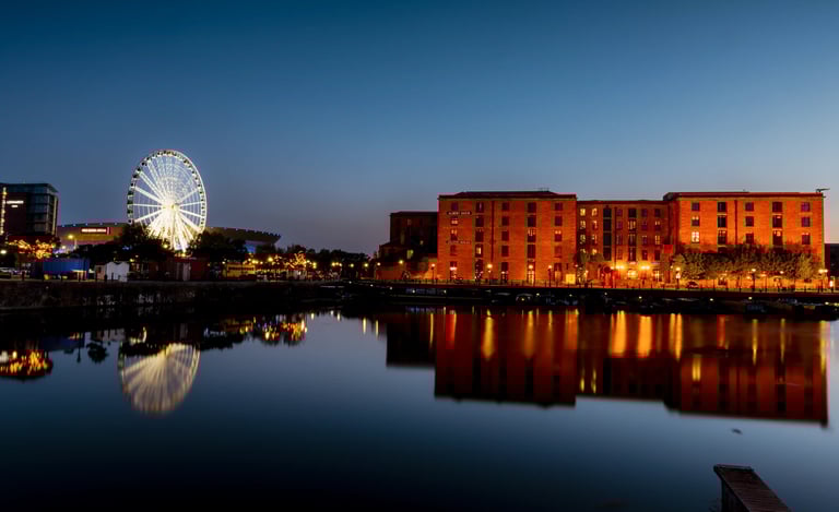 Illuminated ferris wheel and Royal Albert Dock buildings reflected in Liverpool waterfront at night.